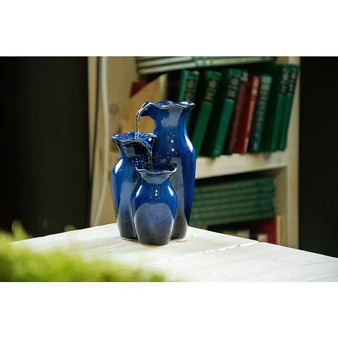 Dobar Zimmerbrunnen - L x B x H: 14 x 12,5 x 22 cm - Nahaufnahme eines dreistufigen blauen Keramikbrunnens auf einem weißen Tisch, mit einem Bücherregal im Hintergrund, der eine beruhigende Atmosphäre schafft.