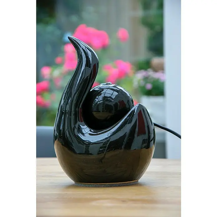 Dobar Zimmerbrunnen Keramik - 16,5 x 14,5 x 23 cm, Keramik - Nahaufnahme einer stilvollen schwarzen Zimmerbrunnen-Dekoration mit einer Kugel und einem geschwungenen Design vor einem floralen Hintergrund.