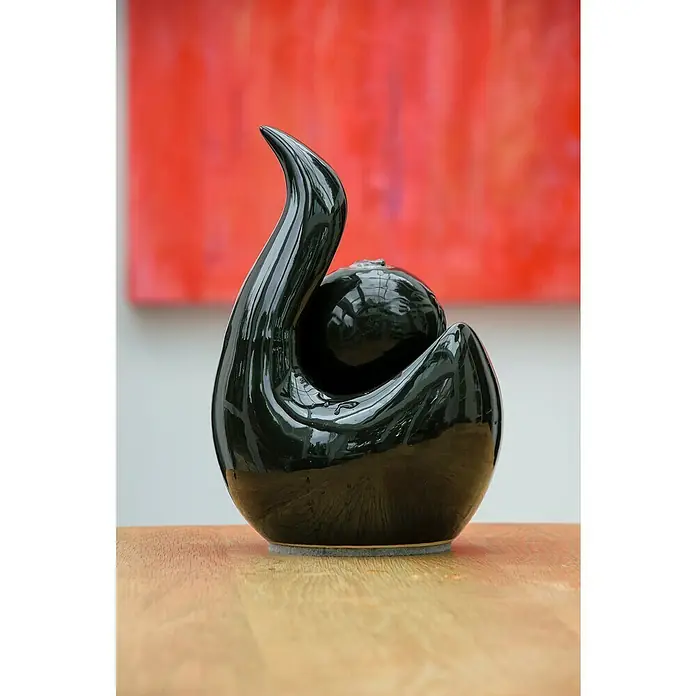Dobar Zimmerbrunnen Keramik - 16,5 x 14,5 x 23 cm, Keramik - Schwarze Keramikskulptur mit geschwungenem Hals und Kugelform, glänzende Oberfläche, steht auf einem Holztisch vor einem roten Hintergrund.