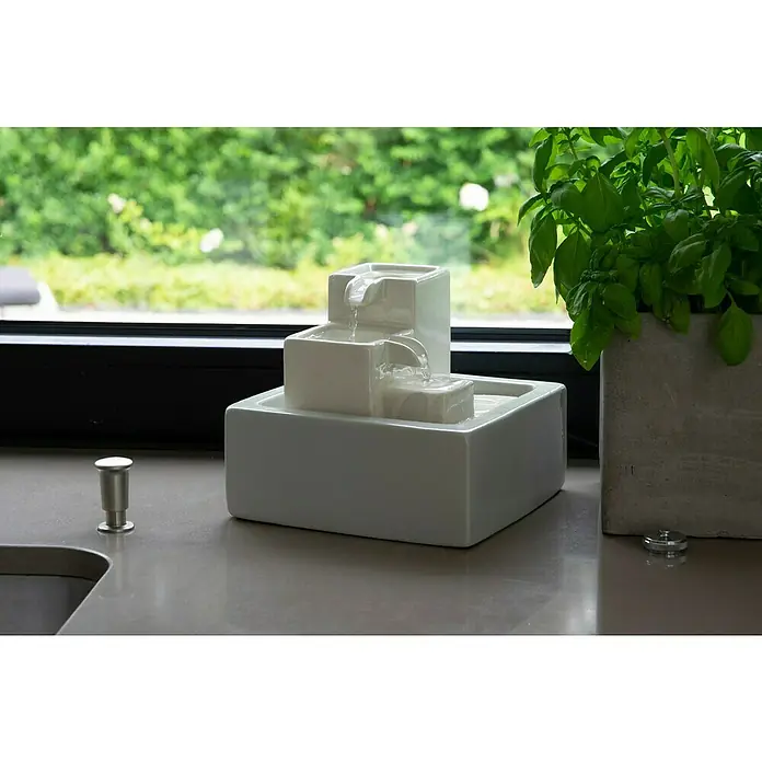 Dobar Zimmerbrunnen - 19 x 19 x 16,5 cm, Keramik - Dekorative Zimmerbrunnen mit drei Ebenen, der neben einem Basilikumtopf auf einer Küchentheke steht.