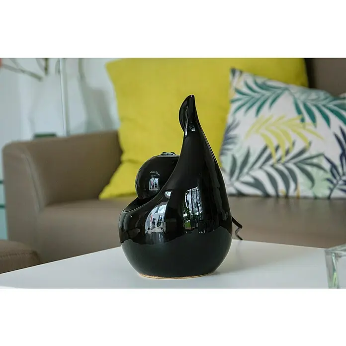 Dobar Zimmerbrunnen Keramik - 16,5 x 14,5 x 23 cm, Keramik - Nahaufnahme einer stilvollen, glänzend schwarzen Zimmerbrunnen-Skulptur auf einem weißen Tisch, vor einem Sofa mit Kissen.