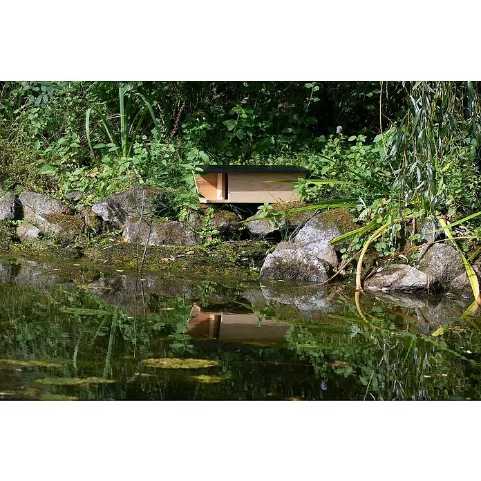 Dobar Nistkasten - 45 x 18 x 15 cm, Natur/ Dunkelbraun, Holz - Holzhaus für Wildtiere mit schwarzem Dach, steht am Ufer eines Teichs, umgeben von Grün. Es spiegelt sich sanft im Wasser.