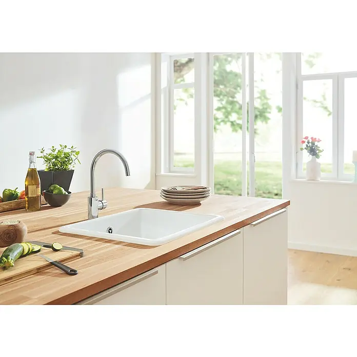 Grohe Concetto Spültischarmatur - C-Auslauf, Chrom, Glänzend, Schwenkbar - Eine moderne Küche mit einer Spüle, einer Armatur, Gemüse und Kräutern auf einer hölzernen Arbeitsplatte vor einem hellen Fenster.