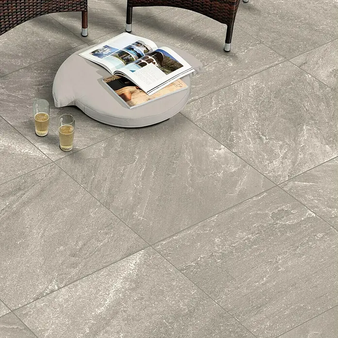 Terrassenfliese Cera 2.0 - 80 x 80 x 2 cm, Quarz Grigio, Matt - Das Bild zeigt eine Nahaufnahme von grauen Keramikfliesen, die eine elegante und moderne Bodenfläche bilden. Ein Buch und zwei Getränke stehen darauf.