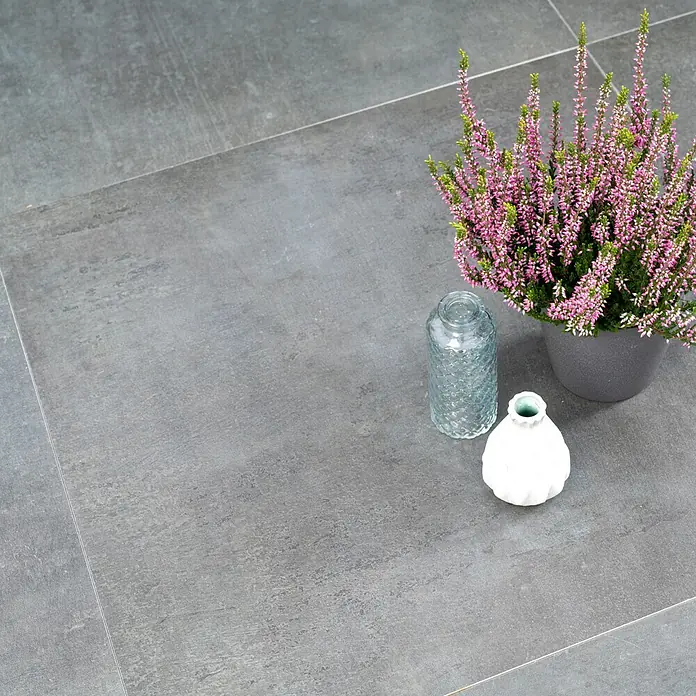 Diephaus Terrassenfliese Columbia - 60 x 60 x 4 cm, Concreto Basalt, Beton - Graue Bodenfliesen mit sanfter Textur bilden die Basis für ein Arrangement aus Heather-Pflanze, Reliefglasvase und einer stilvollen, weißen Dekovase.