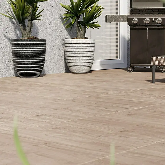 Terrassenfliese Garden Coldbrown 2.0 - 59,7 x 59,7 x 2 cm, Grau/Braun, Matt - Ein Außenbereich mit hellbraunen Bodenfliesen, zwei Pflanzen in gestreiften Töpfen und einem Grill. Die Fliesen verleihen dem Raum eine natürliche Ästhetik.