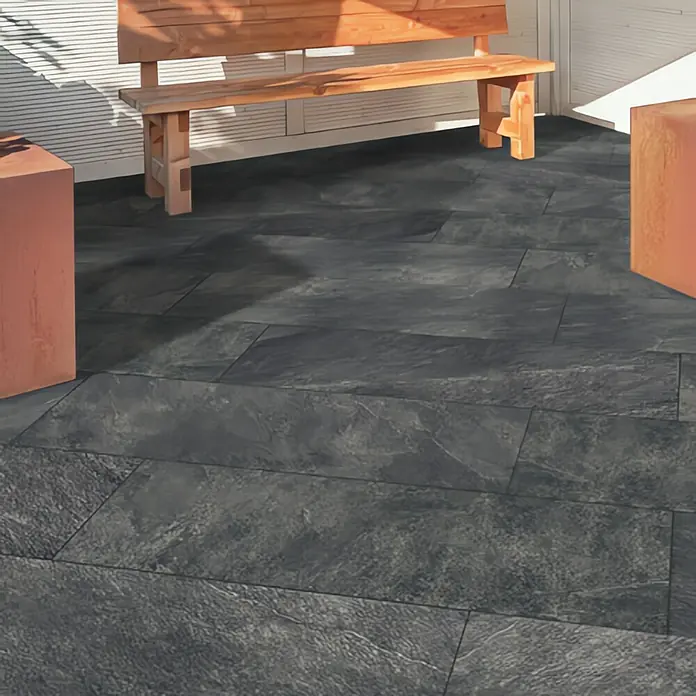 Terrassenfliese Cera 2.0 - 40 x 120 x 2 cm, Museo Ardesia Nero, Matt - Ein Außenbereich mit anthrazitfarbenen Pflastersteinen, einer Holzbank und Pflanzgefäßen, die eine stilvolle Atmosphäre schaffen.