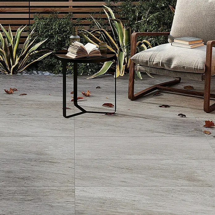 Atlas Concorde Solution Regent Terrassenfliese Silver - 45 x 90 x 2 cm, Silbergrau, Matt - Nahaufnahme von hellgrauen Terrassenfliesen, die eine einladende Außenanlage mit einem Stuhl und einem Tisch mit Büchern zeigen.