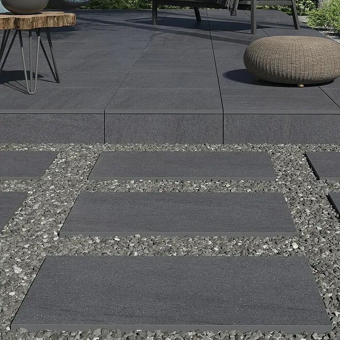 Terrassenfliese Kamari 2.0 - 59,7 x 59,7 x 2 cm, Anthrazit, Matt - Gehwegplatten aus dunklem Stein umgeben von Kies. Perfekt für Gärten, die einen modernen Touch suchen.