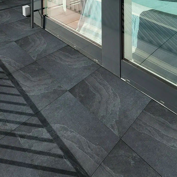 Terrassenfliese Schiefer 2.0 - 60 x 60 x 2 cm, Anthrazit, Matt - Nahaufnahme von dunklen, quadratischen Bodenfliesen mit unregelmäßigem Muster neben einer Glasschiebetür.