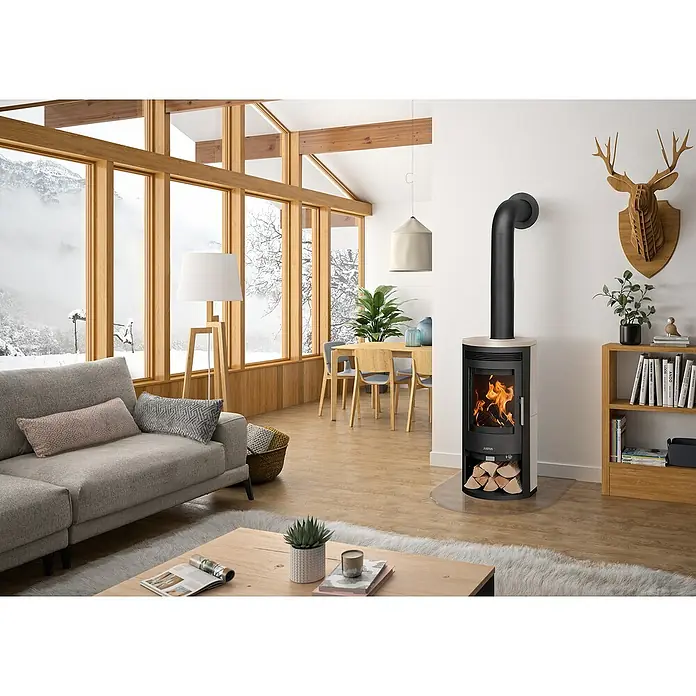 Justus Kaminofen Baltrum D 2.0 - 5,5 kW, Raumheizvermögen: 88 m³, Schwarz/Kalkstein - Kaminofen von Justus mit Holzscheitlagerung und einem schwarzen Rauchrohr in einem hellen Wohnzimmer mit großen Fenstern.