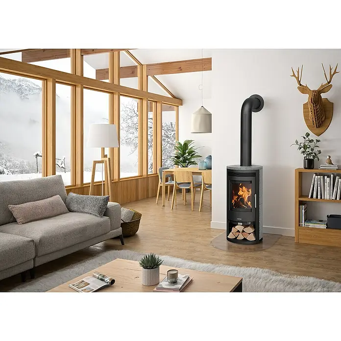 Justus Kaminofen Baltrum D 2.0 - 5,5 kW, Raumheizvermögen: 88 m³, Schwarz/Speckstein - Wohnzimmer mit einem Kaminofen von Justus, der mit Holz befeuert wird, mit Flammen sichtbar hinter der Glasscheibe und Holz darunter gelagert.