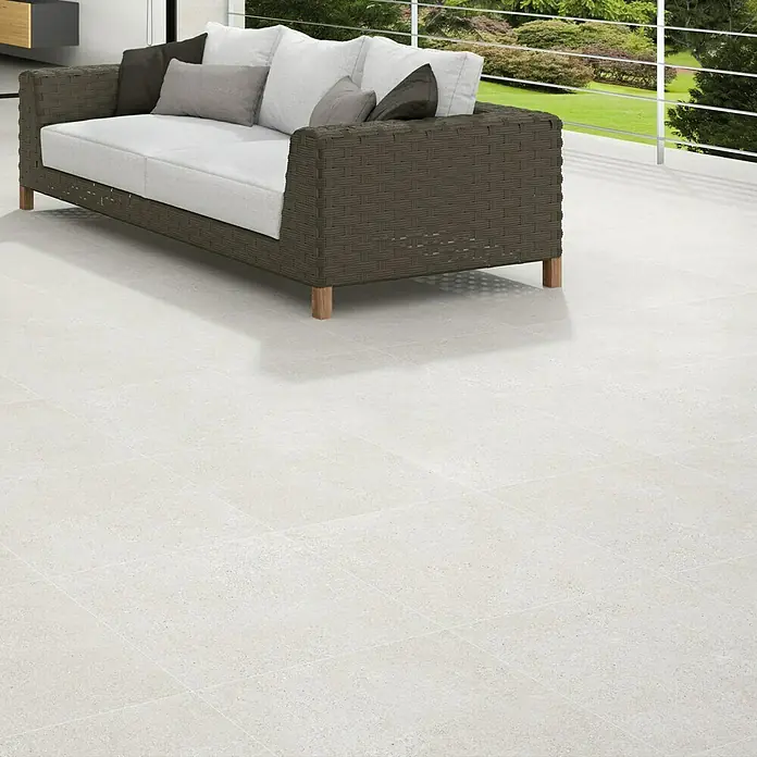 Feinsteinzeugfliese Homestone Grey - 100 x 100 cm, Grau, Matt - Ein graues Rattansofa mit hellen Kissen steht auf einem hellen, gefliesten Boden. Im Hintergrund befindet sich eine Terrasse mit Blick ins Grüne.