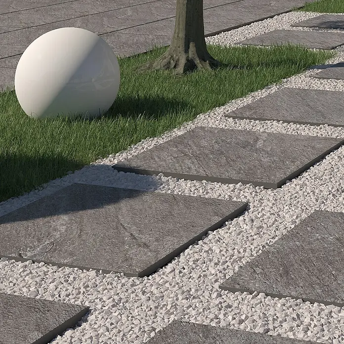 Terrassenfliese Piazza 2.0 - 59,7 x 59,7 x 2 cm, Anthrazit, Matt - Nahaufnahme eines Gartenwegs mit quadratischen Pflastersteinen, umgeben von weißen Kieselsteinen und grünem Gras mit einem Baumstumpf.