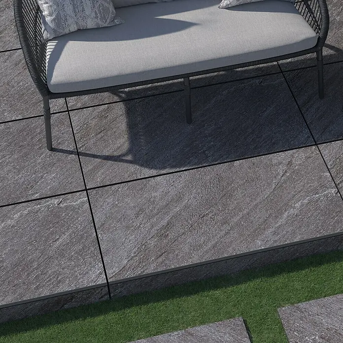 Terrassenfliese Piazza 2.0 - 59,7 x 119,7 x 2 cm, Anthrazit, Matt - Nahaufnahme von grauen Bodenfliesen mit linearen Fugen und einer stilvollen Couch, die eine moderne und elegante Atmosphäre erzeugt.