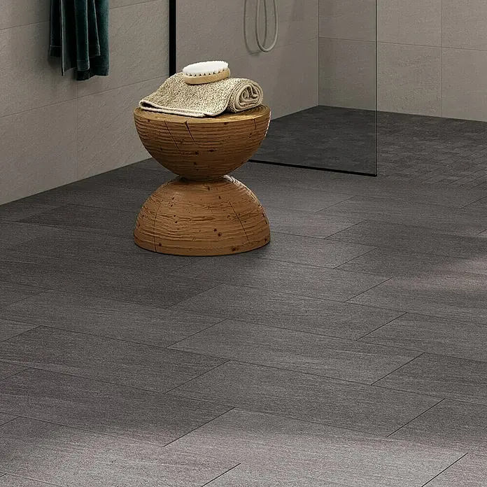 Atlas Concorde Solution Lane Feinsteinzeugfliese - 30 x 60 cm, Asphalt, Matt - Nahaufnahme eines Badezimmerhockers aus Holz mit Handtüchern und Bürste auf einem grauen gefliesten Boden vor einer Dusche.