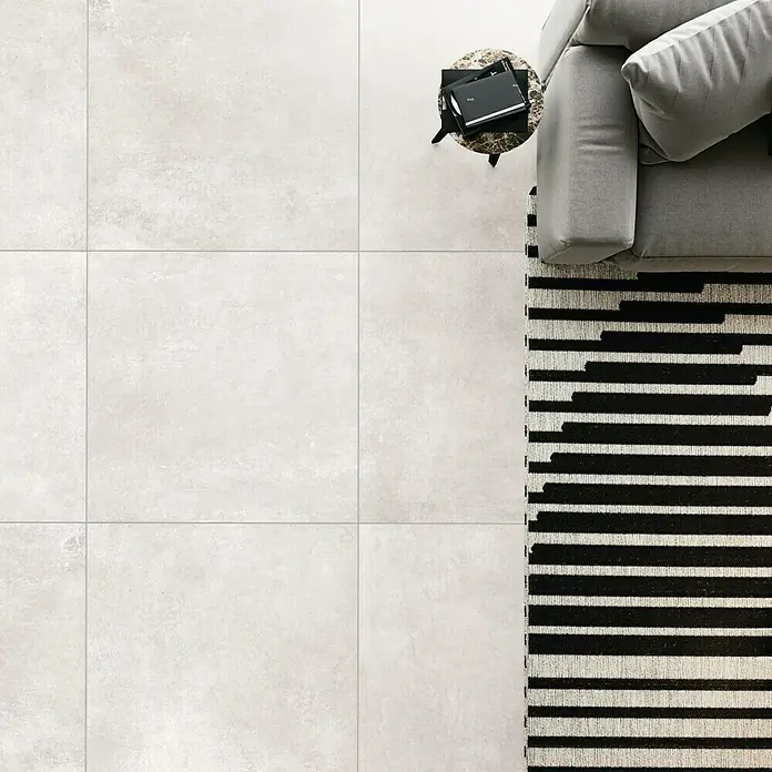 Domicil Feinsteinzeugfliese Living Concrete Pearl - 60 x 60 cm, Beige, Matt - Ein modernes Wohnzimmer mit hellgrauen Fliesen, einem grauen Sofa und einem schwarz-weiß gestreiften Teppich.