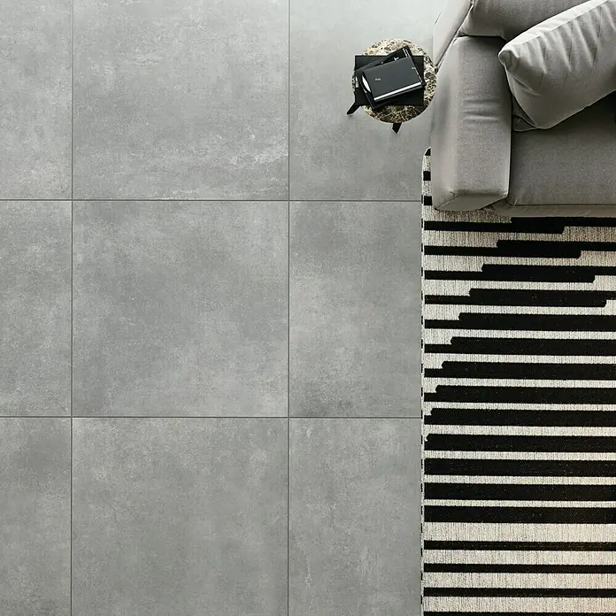 Domicil Feinsteinzeugfliese Living Concrete - 60 x 60 cm, Grau, Matt - Graue Bodenfliesen aus Keramik oder Steinzeug, kombiniert mit einem Sofa und einem Teppich mit Streifenmuster für ein modernes Raumgefühl.