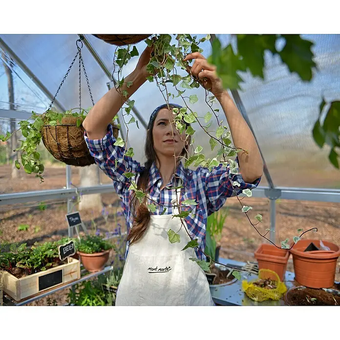 Palram – Canopia Invernadero Hybrid - Medidas ext. (An x Pr): 185 x 248 cm, Plateado, Área: 4,6 m² - Una jardinera de invernadero cuida plantas colgantes, rodeada de una exuberante vegetación en una idílica escena de jardín.