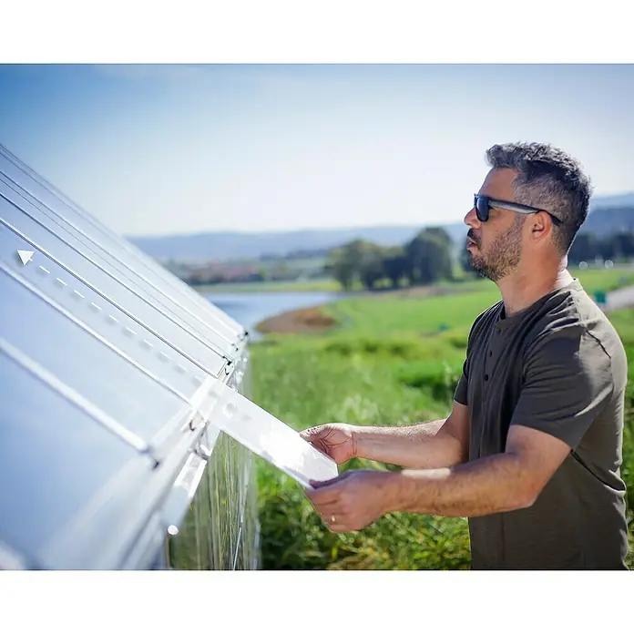 Palram – Canopia Invernadero Balance - Medidas ext. (An x Pr): 304 x 964 cm, 0,7 mm, Plateado - Un hombre instala la cubierta de un invernadero. El panel translúcido se está colocando en la estructura para proteger las plantas.