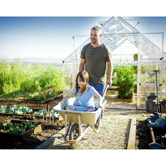 Palram – Canopia Invernadero Balance - Medidas ext. (An x Pr): 304 x 484 cm, 0,7 mm, Plateado - Un padre empuja a su hija en una carretilla por el jardín, con un invernadero al fondo. ¡Diversión en el jardín!