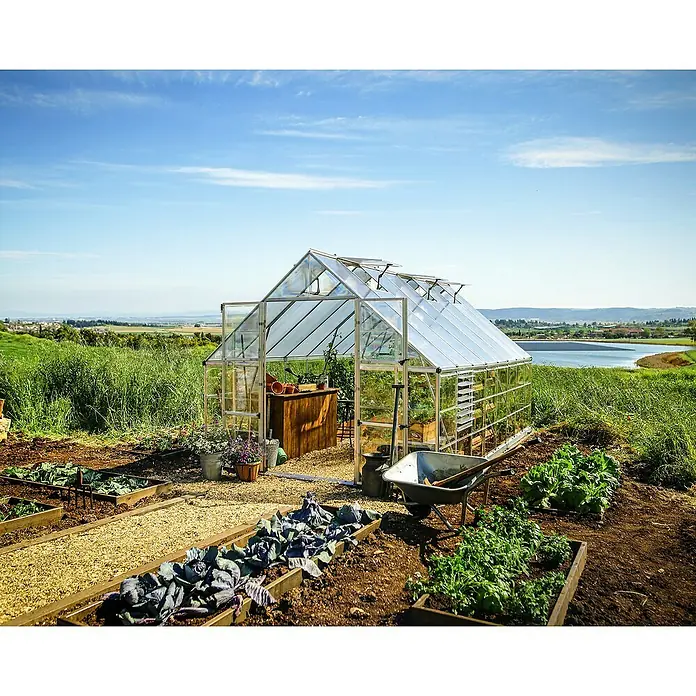 Palram – Canopia Invernadero Balance - Medidas ext. (An x Pr): 304 x 484 cm, 0,7 mm, Plateado - Idílica escena de jardín con invernadero, parcelas de hortalizas y una carretilla frente a un paisaje pintoresco.