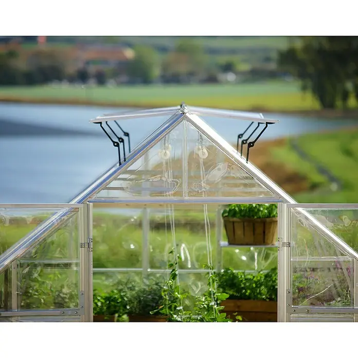 Palram – Canopia Invernadero Balance - Medidas ext. (An x Pr): 304 x 484 cm, 0,7 mm, Plateado - Primer plano de un invernadero en un jardín, con vistas a una serena masa de agua y vegetación exuberante, con plantas que prosperan en su interior.