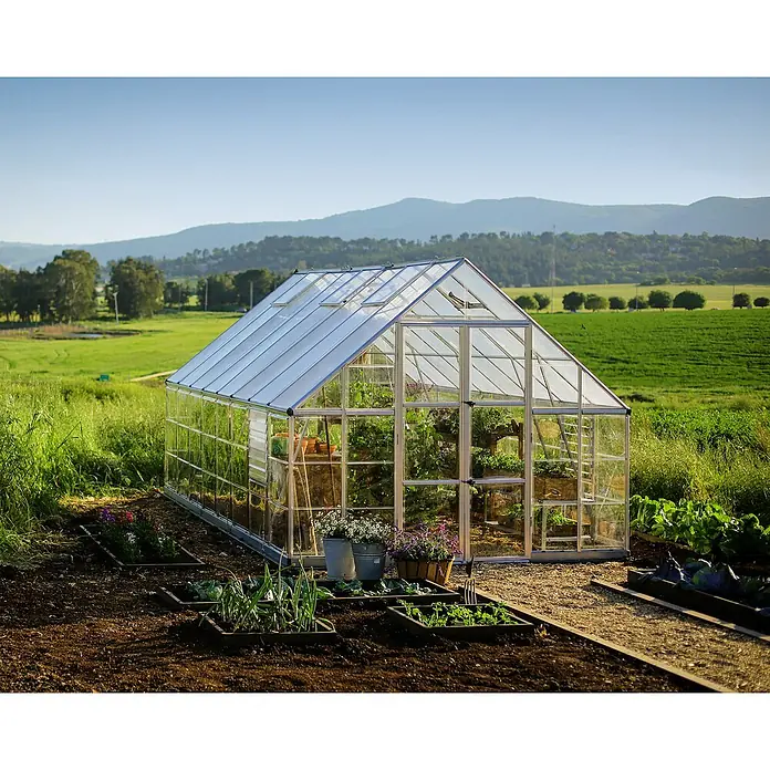 Palram – Canopia Invernadero Balance - Medidas ext. (An x Pr): 304 x 484 cm, 0,7 mm, Plateado - Un invernadero luminoso con plantas y flores frente a un paisaje verde, lo que permite la jardinería.