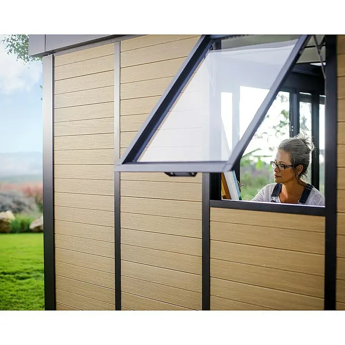 Palram – Canopia Caseta de jardín Copenhaguen - Medidas ext. (An x Pr): 351 x 287 cm, Plástico, Negro - Vista exterior de una caseta de jardín con una ventana abierta, a través de la cual se ve a una mujer pintando. La caseta tiene revestimiento de madera.