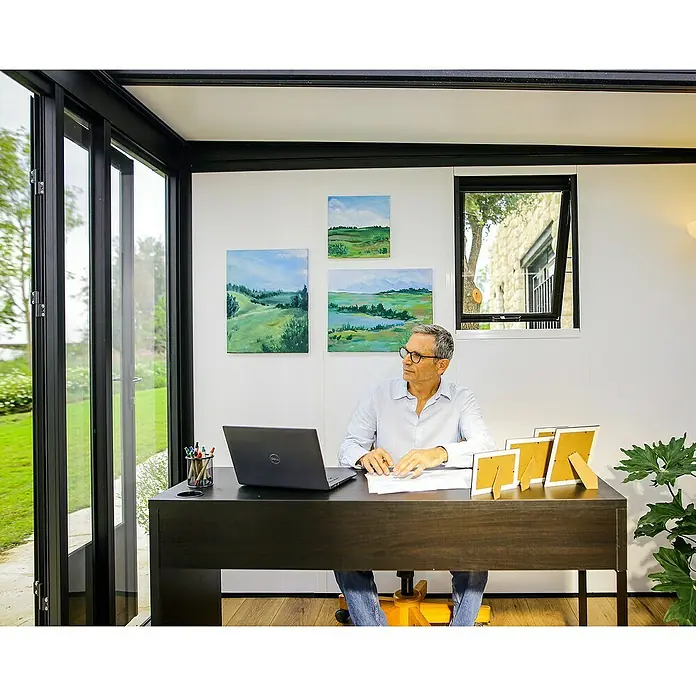 Palram – Canopia Caseta de jardín Copenhaguen - Medidas ext. (An x Pr): 351 x 287 cm, Plástico, Negro - Un hombre está sentado en un escritorio oscuro con un portátil, bolígrafos y marcos de fotos en una oficina moderna con vistas a la vegetación.
