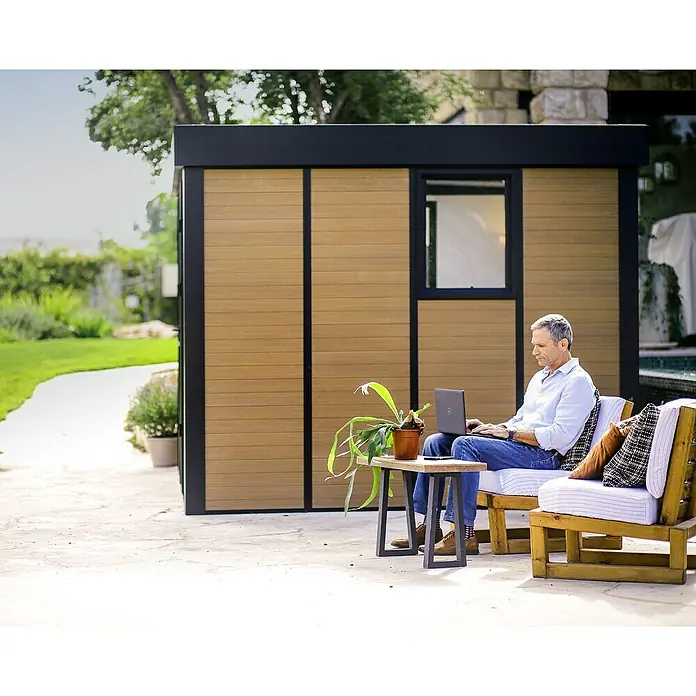 Palram – Canopia Caseta de jardín Copenhaguen - Medidas ext. (An x Pr): 351 x 287 cm, Plástico, Negro - Una caseta de jardín moderna con revestimiento de madera y detalles negros, mientras un hombre está sentado frente trabajando con su portátil.