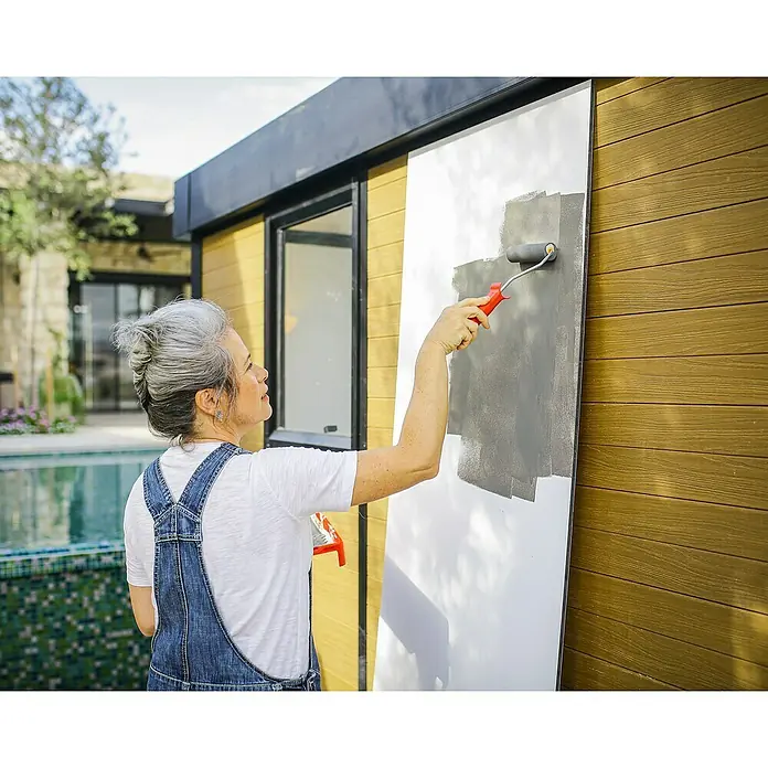 Palram – Canopia Caseta de jardín Copenhaguen - Medidas ext. (An x Pr): 351 x 287 cm, Plástico, Negro - Una mujer está pintando una tabla de gris con un rodillo. La tabla es blanca, se encuentra frente a una casa de madera y una piscina.