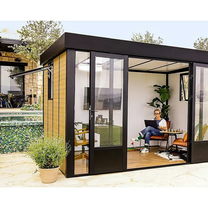 Palram – Canopia Caseta de jardín Copenhaguen - Medidas ext. (An x Pr): 351 x 287 cm, Plástico, Negro - Una moderna oficina de jardín con fachada de vidrio, con una persona trabajando en el interior, ofrece un espacio de trabajo exterior elegante.