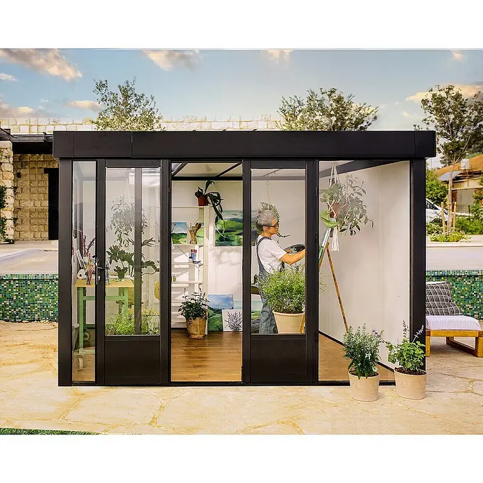 Palram – Canopia Caseta de jardín Copenhaguen - Medidas ext. (An x Pr): 351 x 287 cm, Plástico, Negro - Una artista trabaja dentro de un estudio de jardín moderno y lleno de luz con marcos negros y paredes de cristal. El espacio está decorado con plantas y obras.