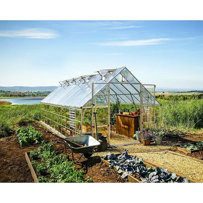 Palram – Canopia Invernadero Balance - Medidas ext. (An x Pr): 304 x 724 cm, 0,7 mm, Plateado - Un espacioso invernadero de cristal con techo abierto se alza en un jardín exuberante con un rico cultivo de hortalizas, con vistas a un lago sereno.