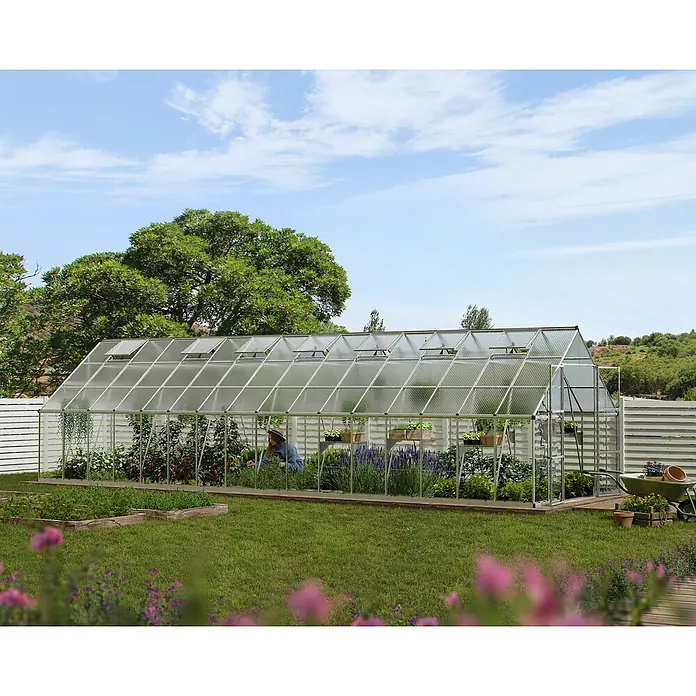 Palram – Canopia Invernadero Balance - Medidas ext. (An x Pr): 304 x 964 cm, 0,7 mm, Plateado - Invernadero con plantas y una persona cuidándolas, situado en un jardín con flores y árboles. Un espacio verde y florido.