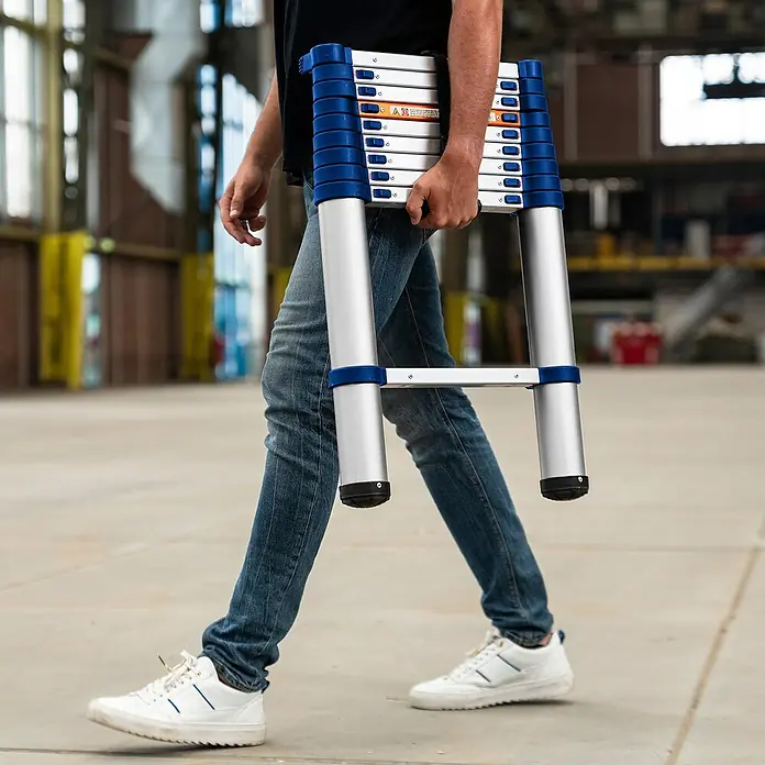 Stabilomat TeleskopleiterArbeitshöhe: 3,63 m, Aluminium - Person, die eine tragbare Teleskopleiter mit blauen Akzenten und silbernen Sprossen durch einen Lagerraum trägt.
