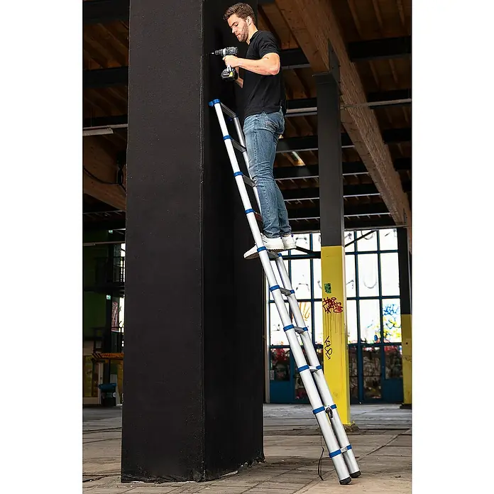 Stabilomat Teleskopleiter - Arbeitshöhe: 3,63 m, Aluminium - Ein Mann steht auf einer Teleskopleiter und bohrt in eine Säule. Die Leiter ist silberfarben mit blauen Akzenten und steht in einem großen Raum.
