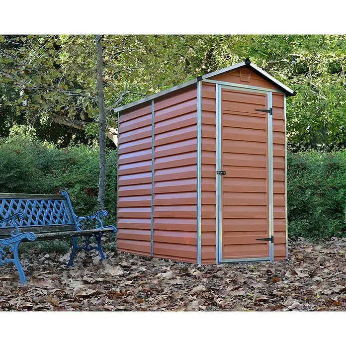 Palram – Canopia Caseta de jardín Skylight - Medidas ext. (An x Pr): 121 x 176 cm, Plástico, Ámbar - Una caseta de jardín marrón con puerta está al lado de un banco de parque azul, rodeada de árboles y hojas.