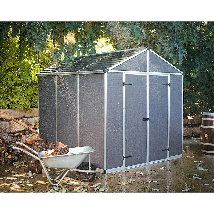 Palram – Canopia Caseta de jardín Rubicon - Medidas ext. (An x Pr): 237 x 229 cm, Plástico, Antracita - Una caseta de jardín gris espaciosa se alza en el jardín, junto a ella una carretilla con leña y una barrica de vino al fondo.