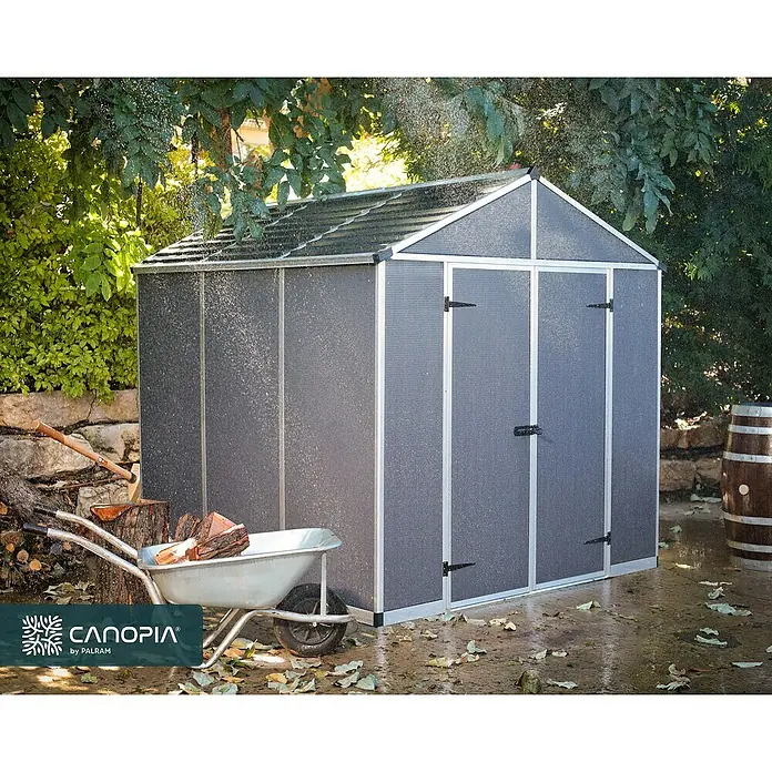 Palram – Canopia Caseta de jardín Rubicon - Medidas ext. (An x Pr): 237 x 229 cm, Plástico, Antracita - Un espacioso cobertizo gris Canopia by Palram, rodeado de exuberante vegetación y herramientas de jardinería. Perfecto para almacenamiento.
