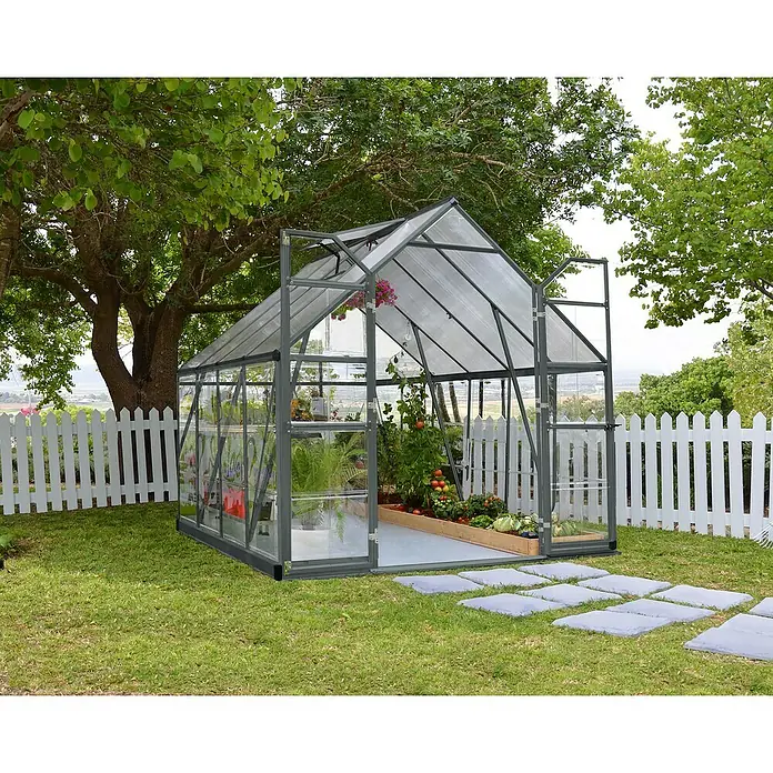 Palram – Canopia Invernadero Balance - Medidas ext. (An x Pr): 244 x 247 cm, 0,7 mm, Gris - Un espacioso invernadero de metal y vidrio se encuentra en el jardín, rodeado por una cerca blanca y exuberante vegetación.