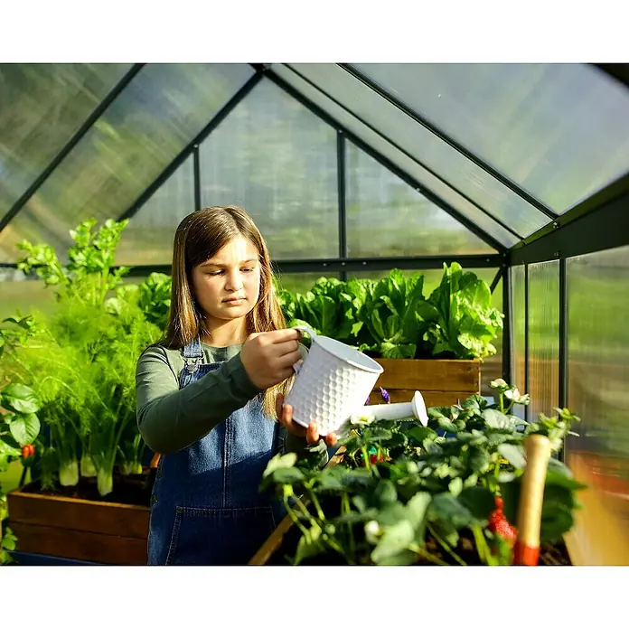 Palram – Canopia Invernadero Mythos - Medidas ext. (An x Pr): 185 x 306 cm, 4 mm, Gris - Una niña riega plantas en un invernadero con una regadera blanca, destacando la jardinería y el cuidado de las plantas.
