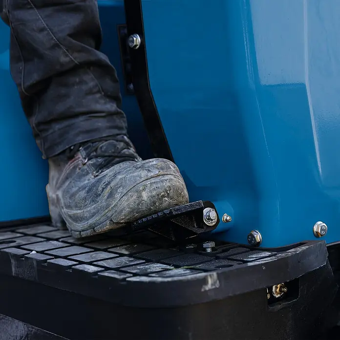 Scheppach Minibagger EXC815 - 6,3 kW, Auslegerschwenkbereich: -70 ° - 70 °, Benzin, Blau - Fuß auf Trittbrett eines Arbeitsfahrzeugs. Der robuste Stiefel betont die Strapazierfähigkeit des Geräts.