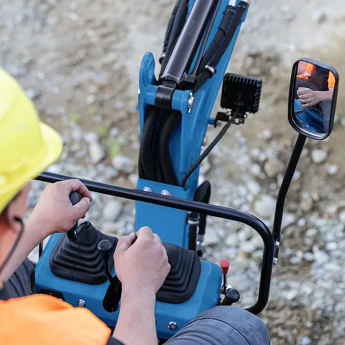 Scheppach Minibagger EXC815 - 6,3 kW, Auslegerschwenkbereich: -70 ° - 70 °, Benzin, Blau - Nahaufnahme eines Bauarbeiters mit gelbem Helm, der einen blauen Minibagger bedient. Fokus auf Bedienelemente und Spiegel.