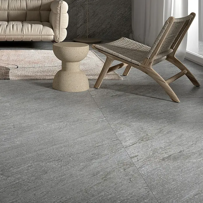 Feinsteinzeugfliese Piazza Grey - 59,7 x 59,7 cm, Grau, Matt - Nahaufnahme eines stilvollen Innenraums mit grauen Bodenfliesen, einem bequemen Sofa, einem Beistelltisch und einem modernen Stuhl.