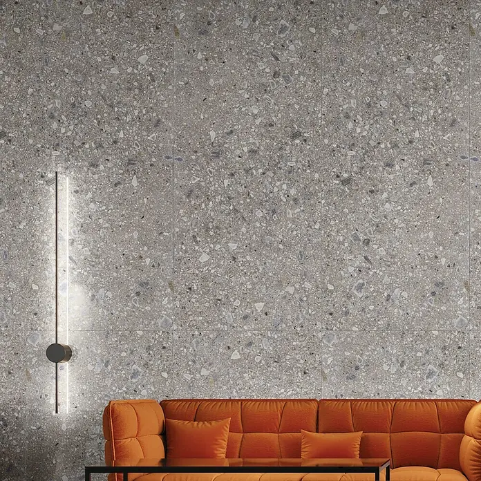 Feinsteinzeugfliese Terrazzo Ceppo119,7 x 119,7 cm, Silber, Glänzend - Wandfliesen in Terrazzo-Optik mit einer modernen Wandleuchte und einem orangefarbenen Sofa.