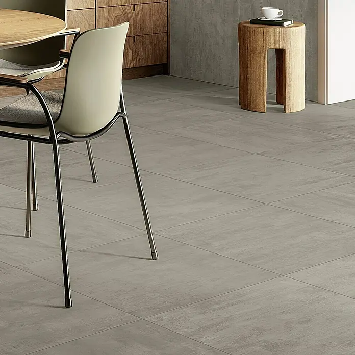 Atlas Concorde Solution Yorkshire Feinsteinzeugfliese Ash - 60 x 60 cm, Grau, Matt - Moderner Innenraum mit grauen Bodenfliesen, Stuhl und Holzhocker mit Tasse. Elegantes Fliesendesign für ein stilvolles Zuhause.