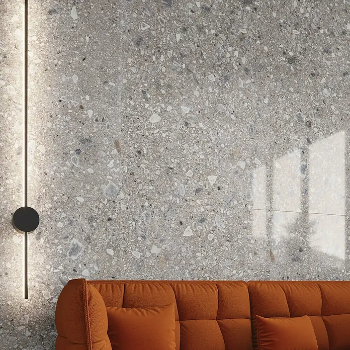 Feinsteinzeugfliese Terrazzo Ceppo - 59,7 x 59,7 cm, Silber, Glänzend - Wandfliesen in Terrazzo-Optik mit orangenem Sofa. Die Fliesen haben ein Muster aus kleinen Steinen und reflektieren das Licht.