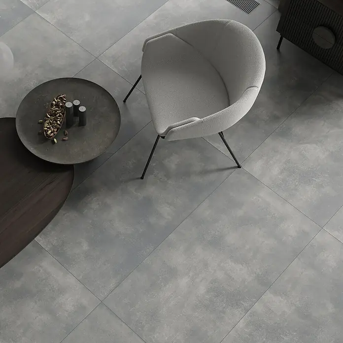 Feinsteinzeugfliese Big Show XXL - 59,8 x 119,8 cm, Grafit, Matt - Elegantes Interieur mit einem hellgrauen Sessel, stilvollen Couchtischen und modernem Fliesenboden, der eine ruhige Atmosphäre schafft.